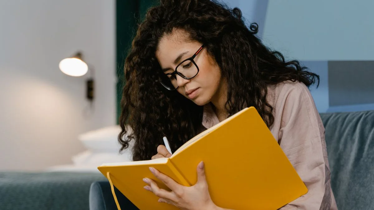 A person writing in a notebook on a sofa beside a warm lamp