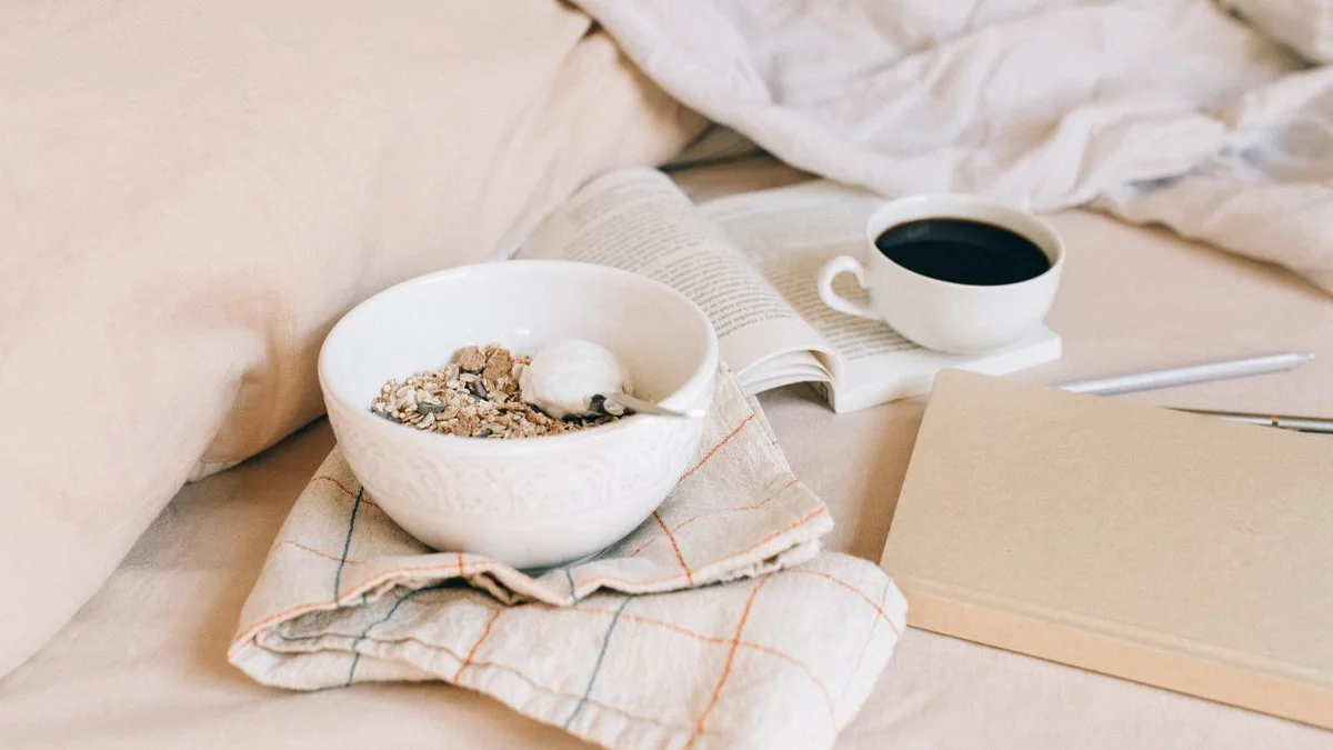 Breakfast, a book and a plain notebook with a pen on a bed