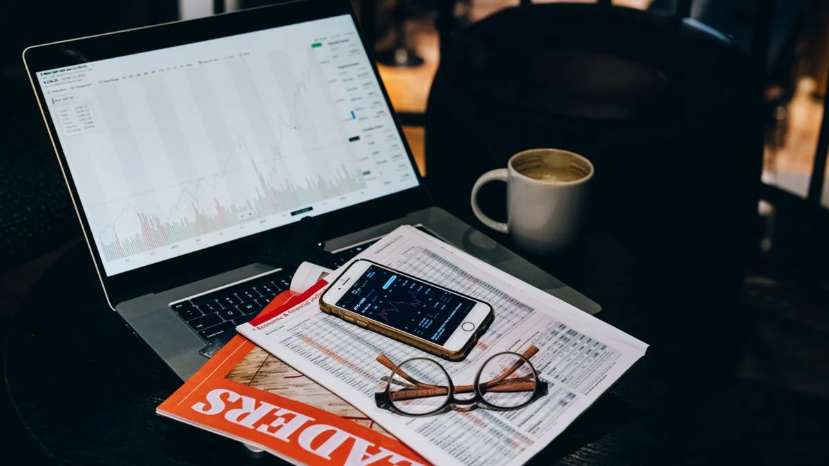 A laptop showing a chart next to a phone, printed financial tables and reading glasses