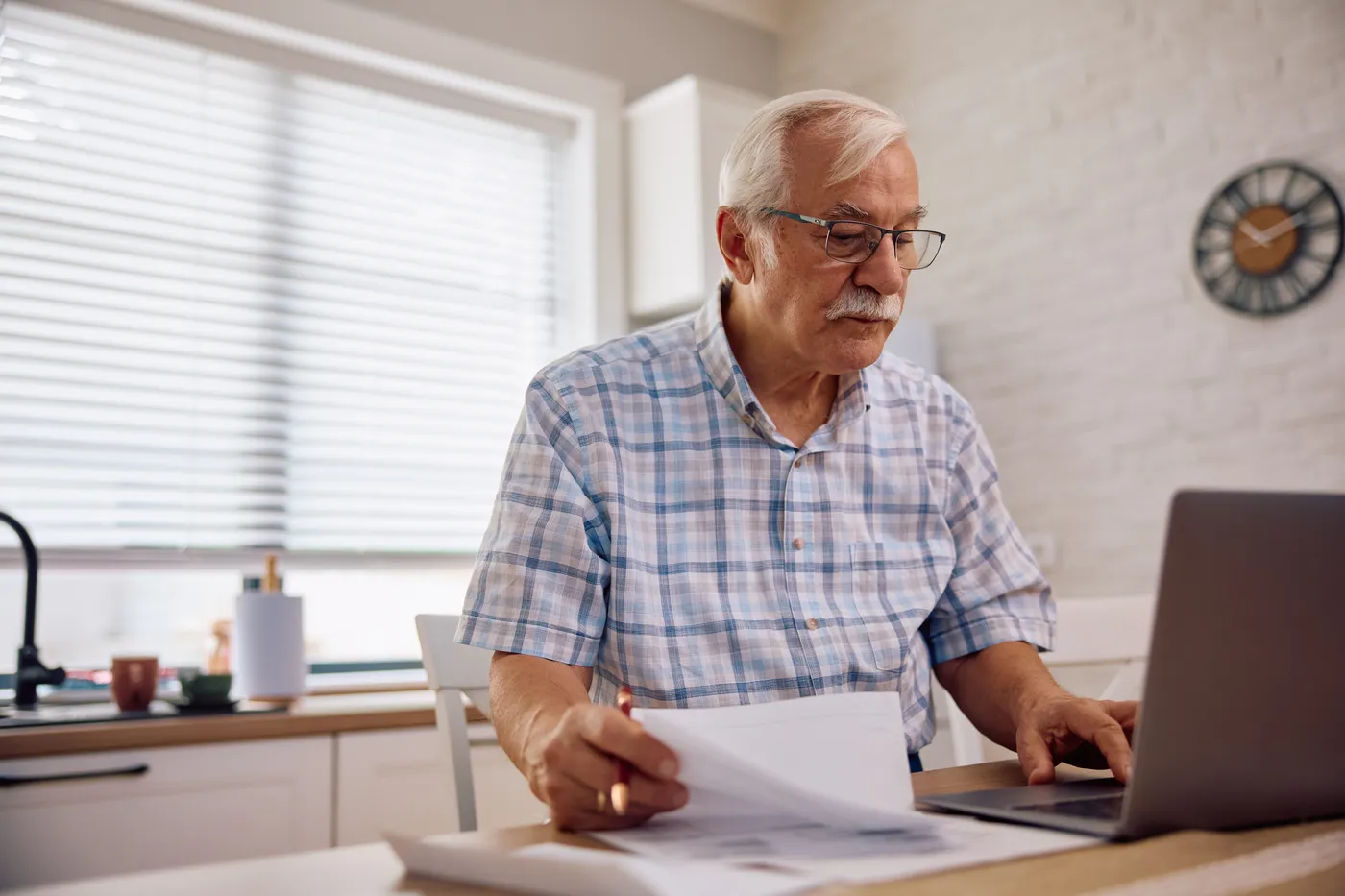 An older man at his kitchen table checks a printed statement against his laptop